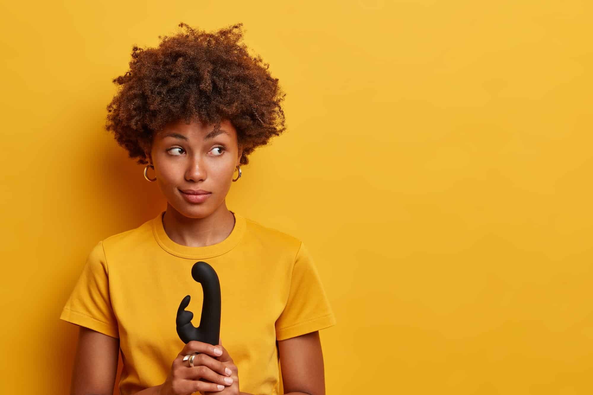 Pensive beautiful woman with Afro hair going to explore vibrating power of new sex toy, holds vibrat